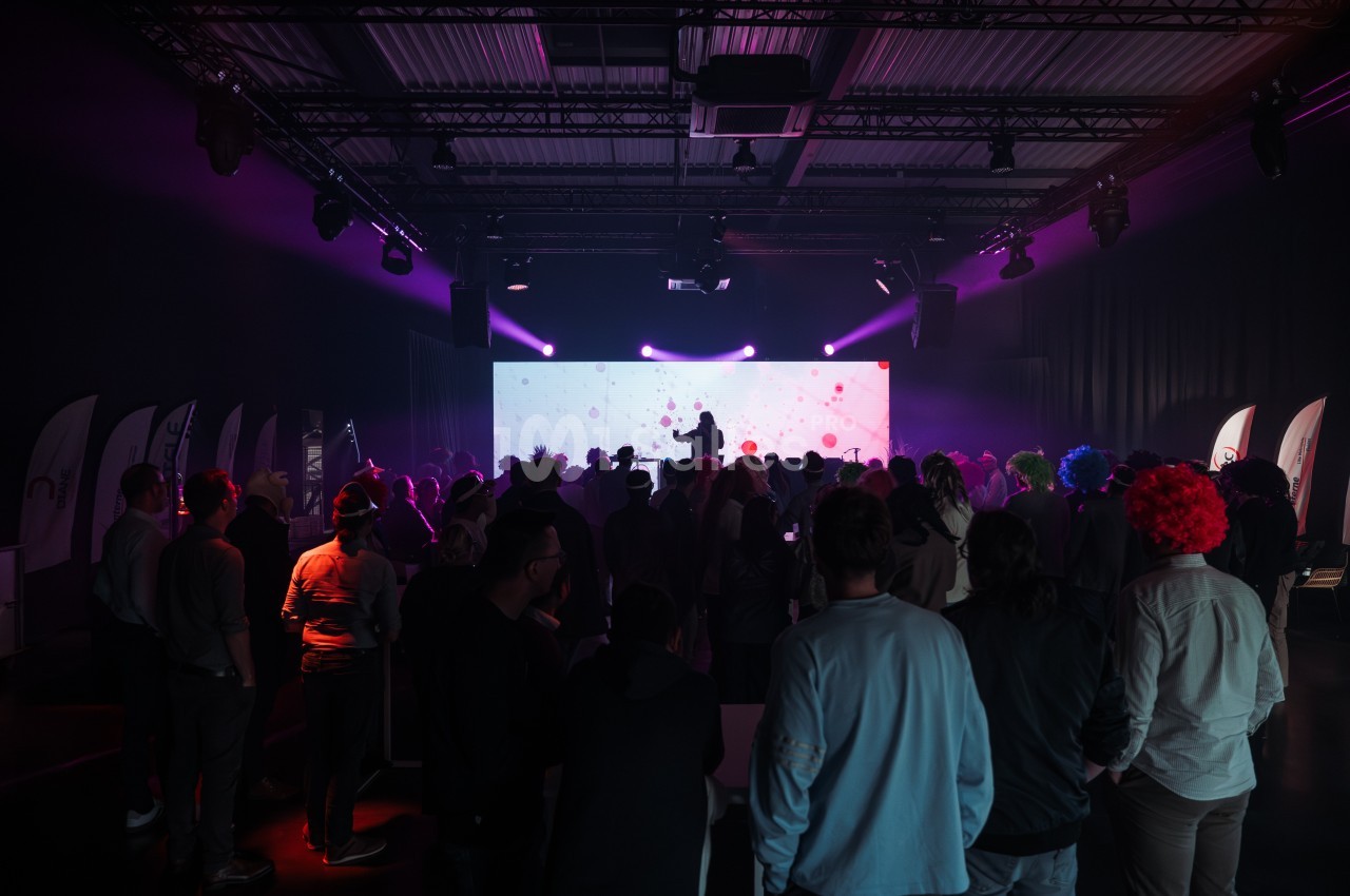 Une foule assiste à une présentation sur scène dans une salle sombre éclairée par des lumières colorées.