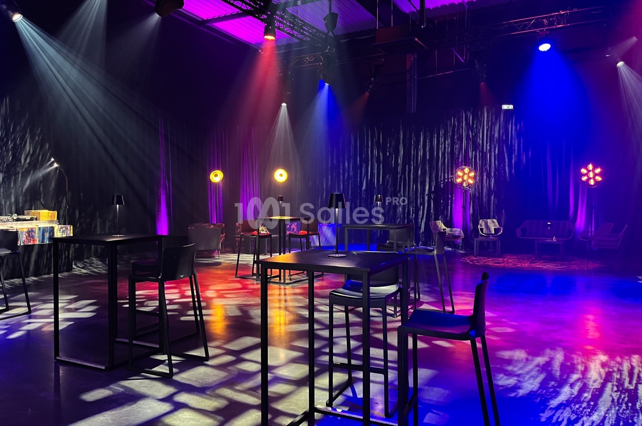 Salle sombre aménagée avec des tables hautes, des chaises et des éclairages colorés créant une ambiance festive.