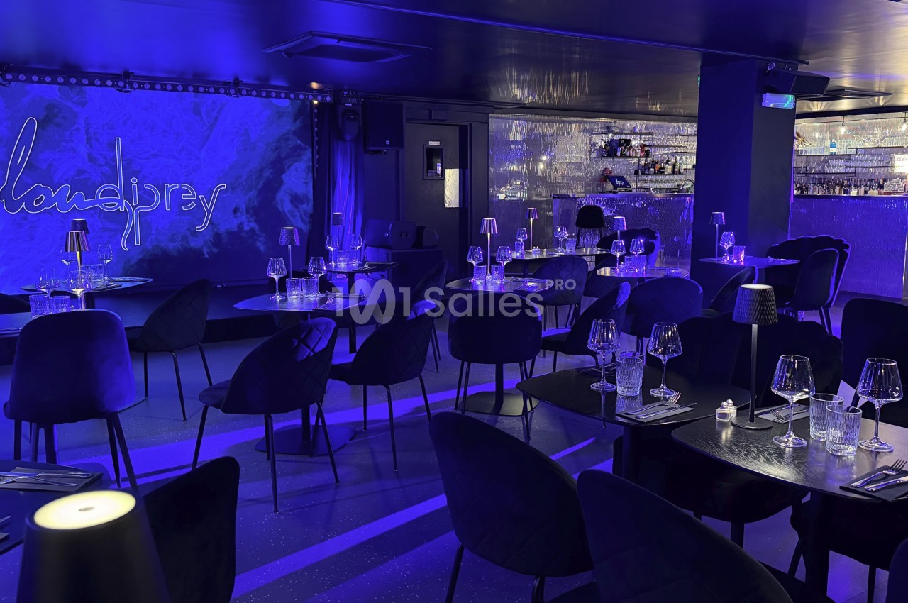 Salle de restaurant moderne avec éclairage bleu, tables dressées et un bar illuminé en arrière-plan.