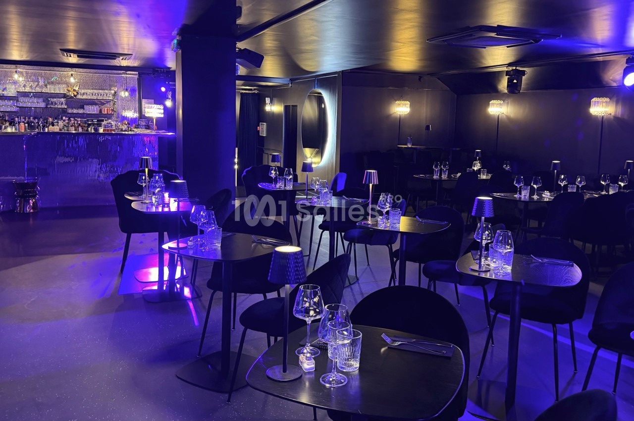 Salle de restaurant élégante avec éclairage tamisé violet, tables dressées et bar en arrière-plan.