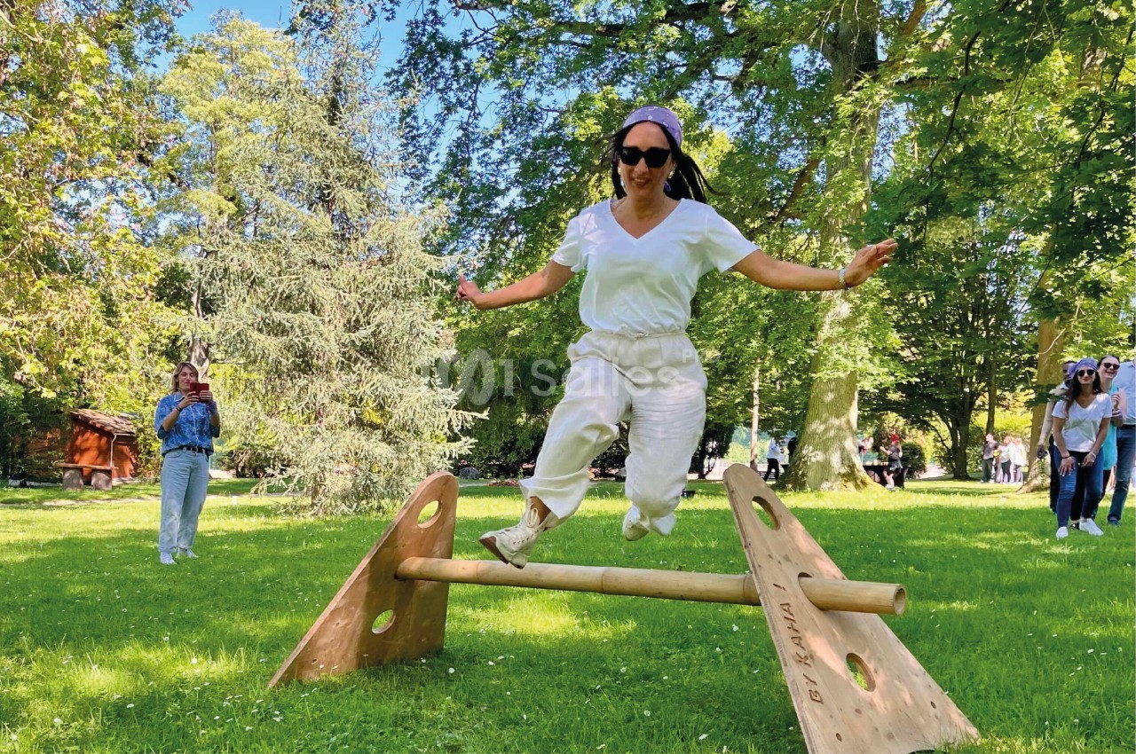 Une femme saute par-dessus une barre en bois dans un parc verdoyant, sous un ciel ensoleillé.