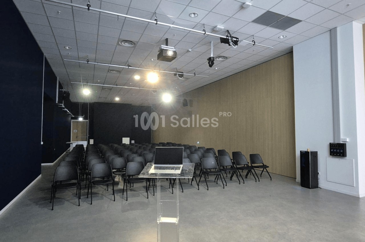 Salle de conférence vide avec des rangées de chaises noires, un pupitre transparent et des projecteurs allumés.