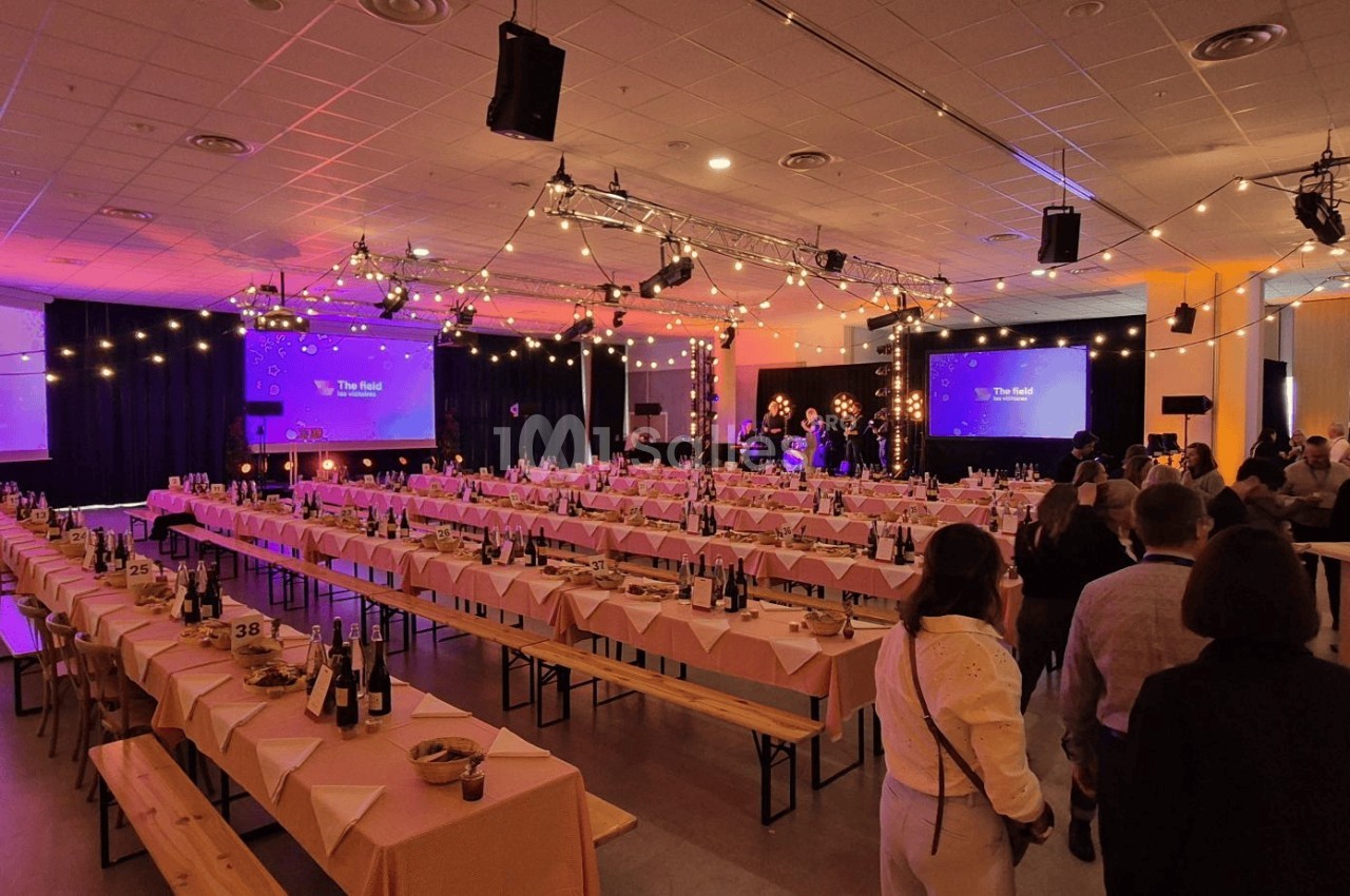Salle de réception décorée avec des guirlandes lumineuses, tables dressées et scène en arrière-plan.