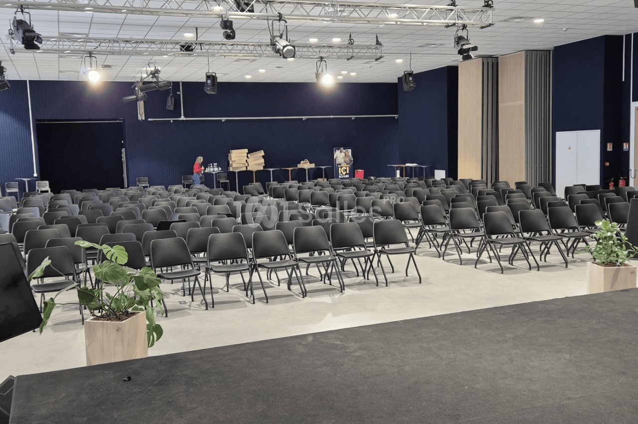 Salle de conférence avec des rangées de chaises noires alignées, vue depuis une scène équipée d'écrans et de plantes.