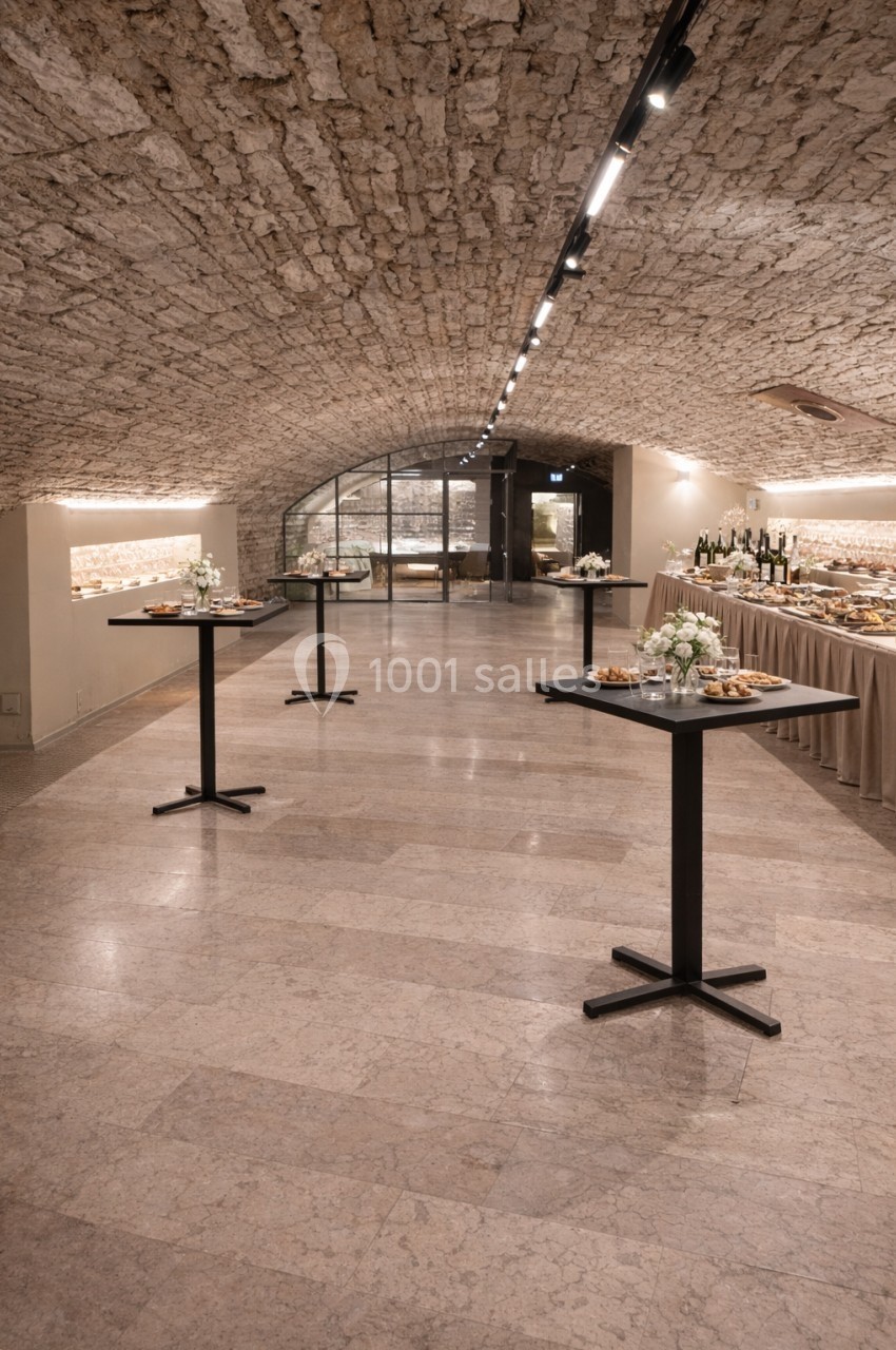 Salle voûtée en pierre avec tables hautes dressées et buffet disposé sur le côté.