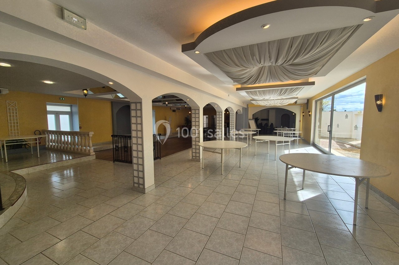 Salle spacieuse avec carrelage clair, arches décoratives, tables rondes et lumière naturelle provenant de grandes baies…