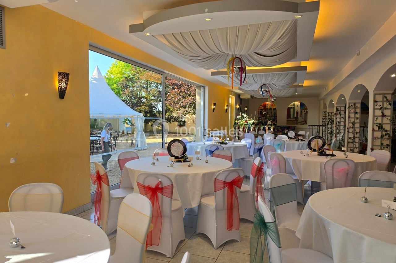 Salle de réception lumineuse avec tables rondes décorées, chaises ornées de nœuds colorés et vue sur un jardin extérieur.