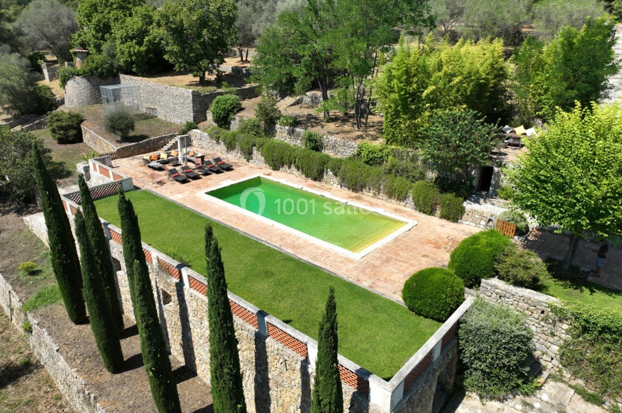 Vue aérienne d'une piscine rectangulaire entourée de végétation, avec des transats sur une terrasse en pierre.