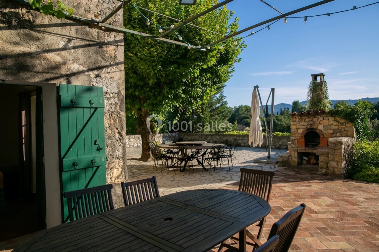 Terrasse ensoleillée avec table en bois, barbecue en pierre, arbres et vue sur un paysage vallonné.