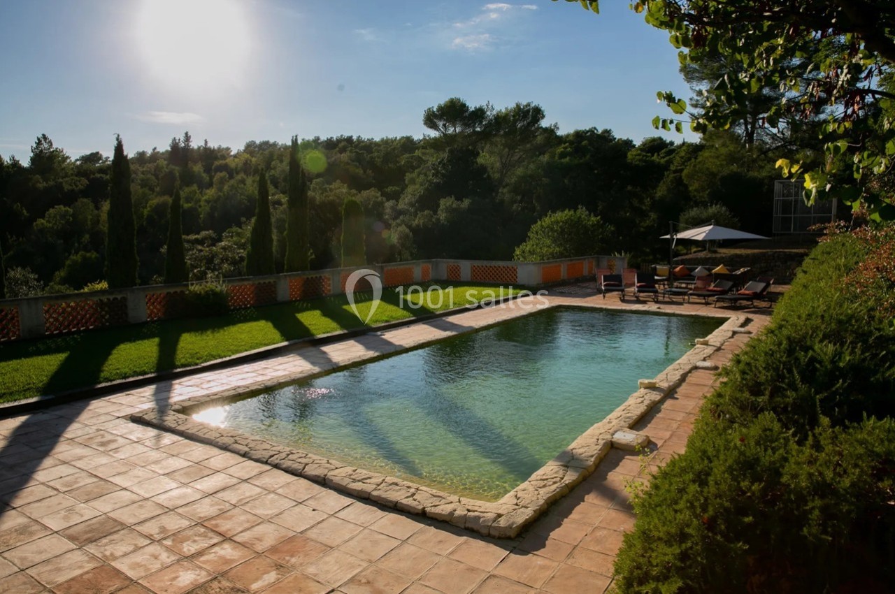 Piscine extérieure entourée de végétation, avec terrasse en pierre et chaises longues sous un ciel ensoleillé.