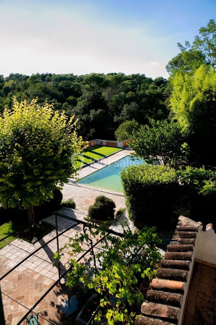 Vue d'une piscine entourée de végétation luxuriante, avec une terrasse en briques au premier plan.