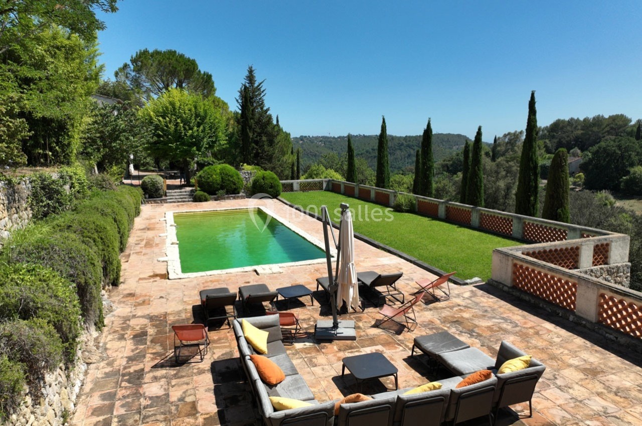 Terrasse en pierre avec piscine, chaises longues et canapés, entourée de verdure et offrant une vue sur la campagne.