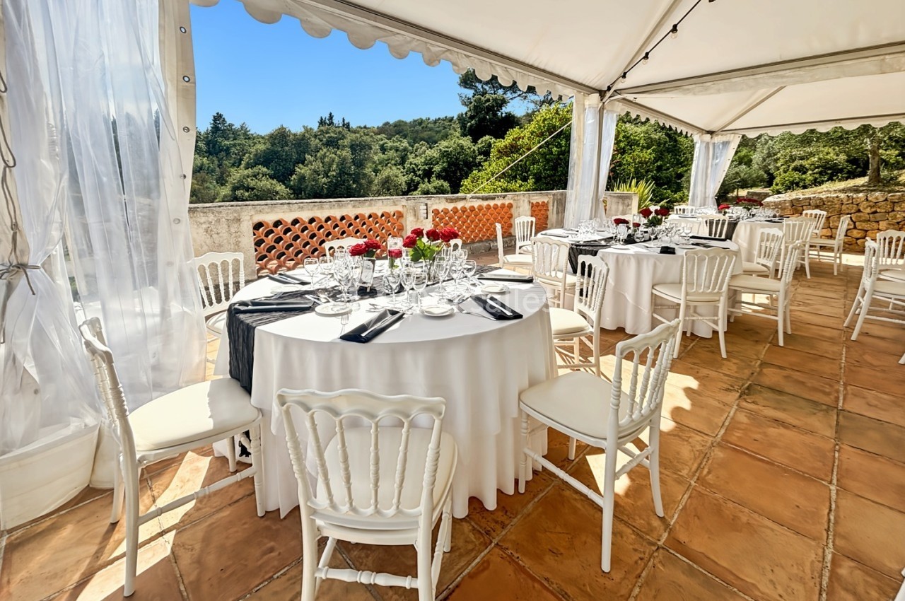 Tables rondes dressées avec nappes blanches et chaises élégantes sous une tente ouverte, donnant sur un paysage verdoyant.