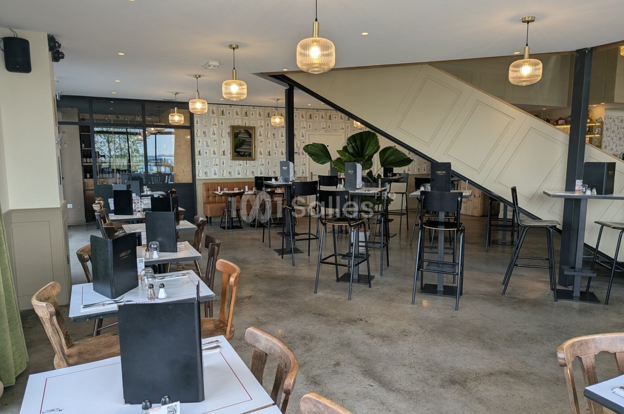 Salle de restaurant moderne avec tables dressées, chaises hautes, escalier central et décoration lumineuse.