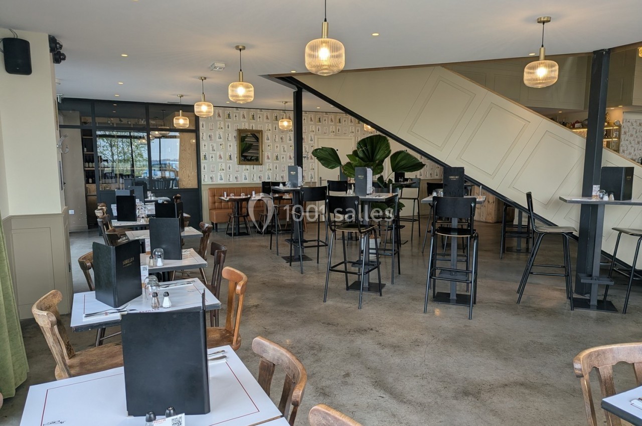 Salle de restaurant moderne avec tables dressées, chaises hautes, escalier et grandes plantes décoratives.