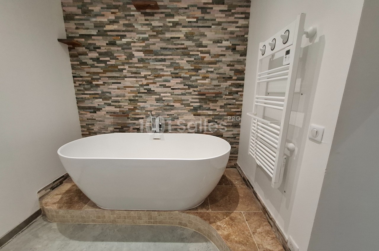 Baignoire blanche sur une plateforme carrelée, devant un mur en pierre naturelle avec un sèche-serviettes à droite.