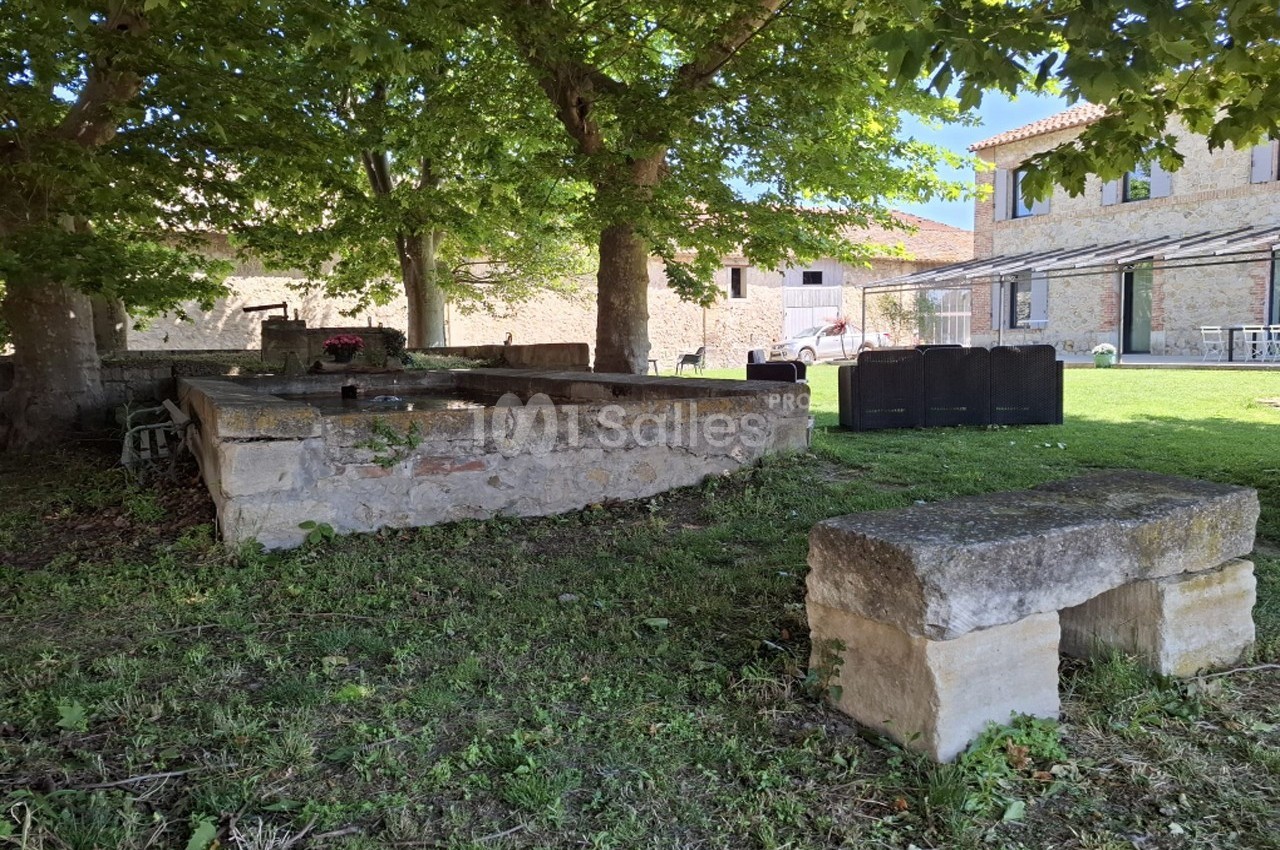 Banc en pierre et ancien lavoir sous des arbres, entourés de verdure, près d'un bâtiment en pierre.