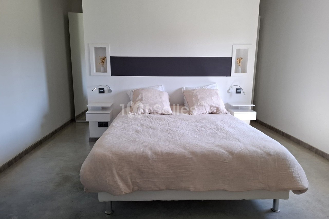 Lit double avec draps beiges dans une chambre minimaliste aux murs clairs et sol en béton.