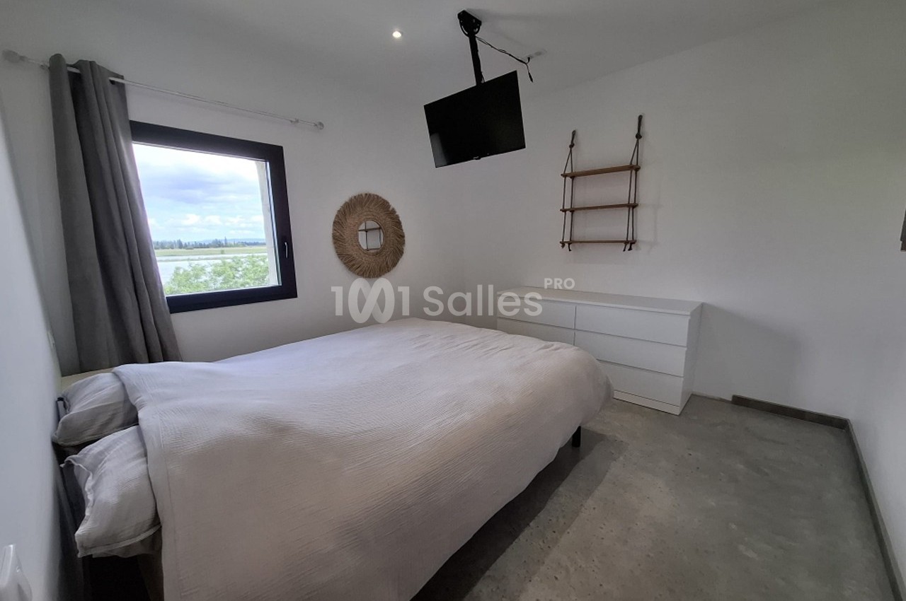 Chambre lumineuse avec lit double, commode blanche, étagères murales, fenêtre donnant sur un paysage et télévision suspendue.