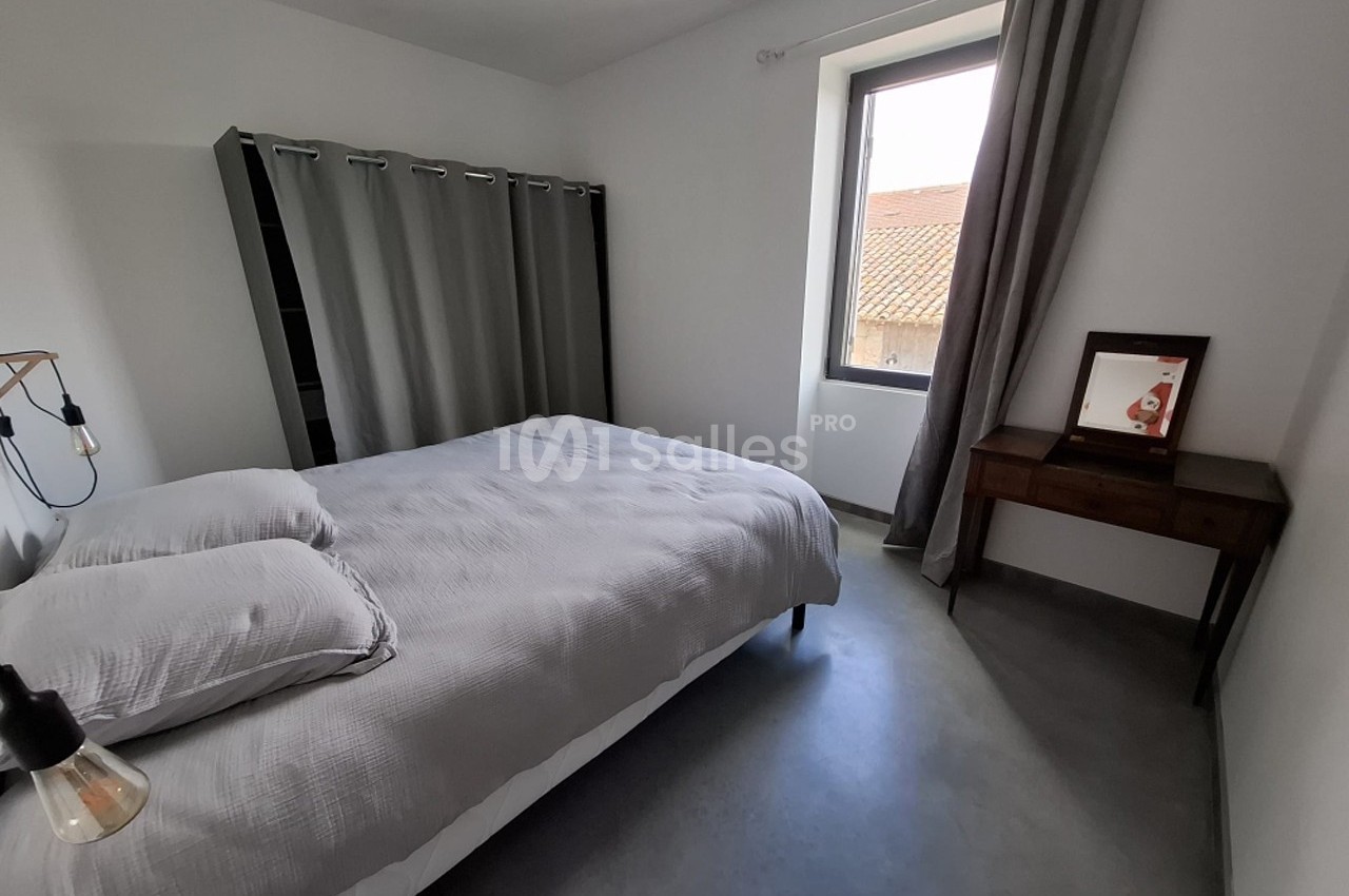 Chambre minimaliste avec lit double, rideaux gris, bureau en bois et fenêtre donnant sur un toit en tuiles.