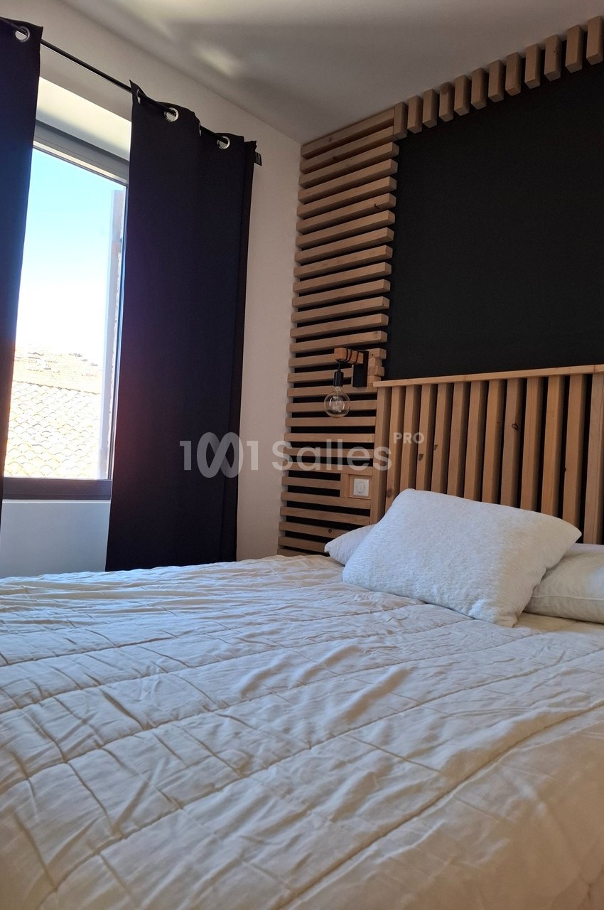 Lit double avec couverture claire, tête de lit en bois, lampe murale et fenêtre avec rideaux noirs laissant entrer la…