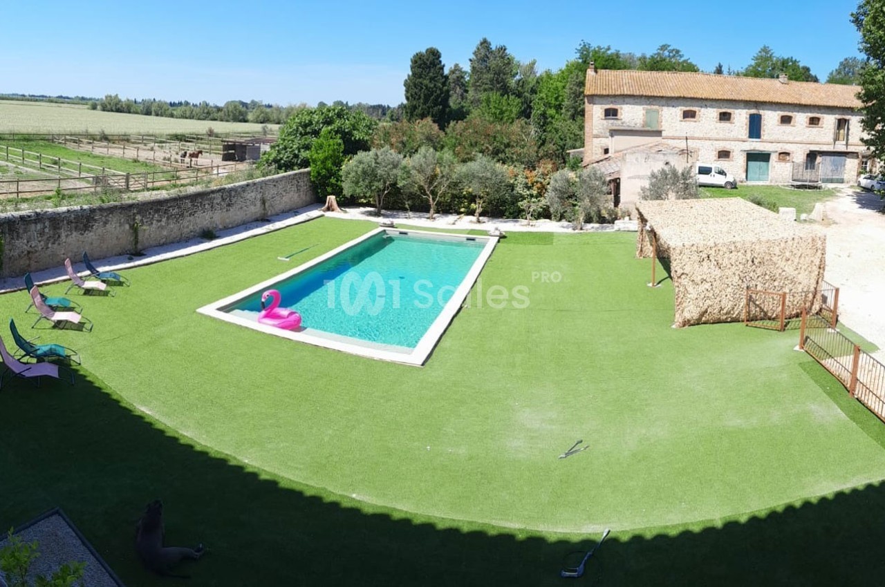 Vue d'une piscine extérieure entourée de gazon artificiel, avec des chaises longues, une bouée rose et des bâtiments en…