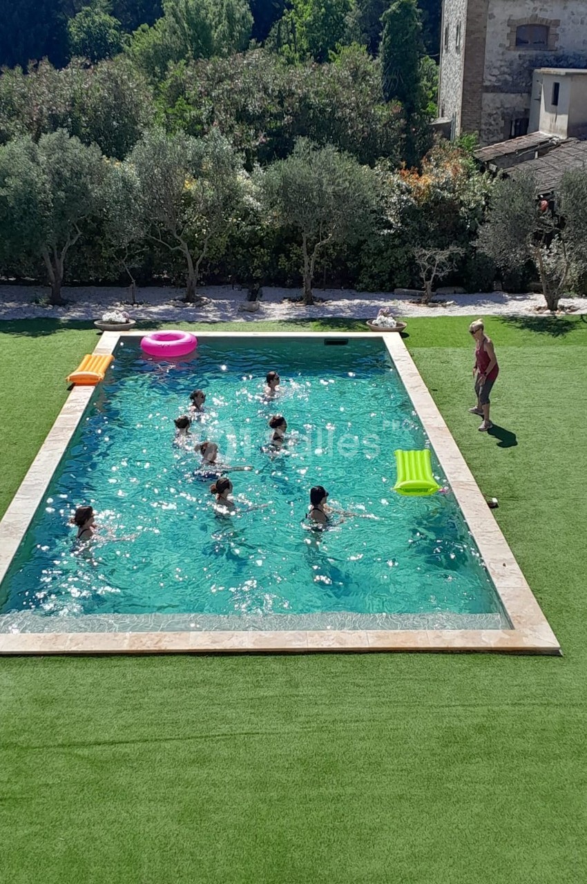 Vue aérienne d'une piscine extérieure entourée de pelouse, avec plusieurs personnes nageant et un homme debout au bord.