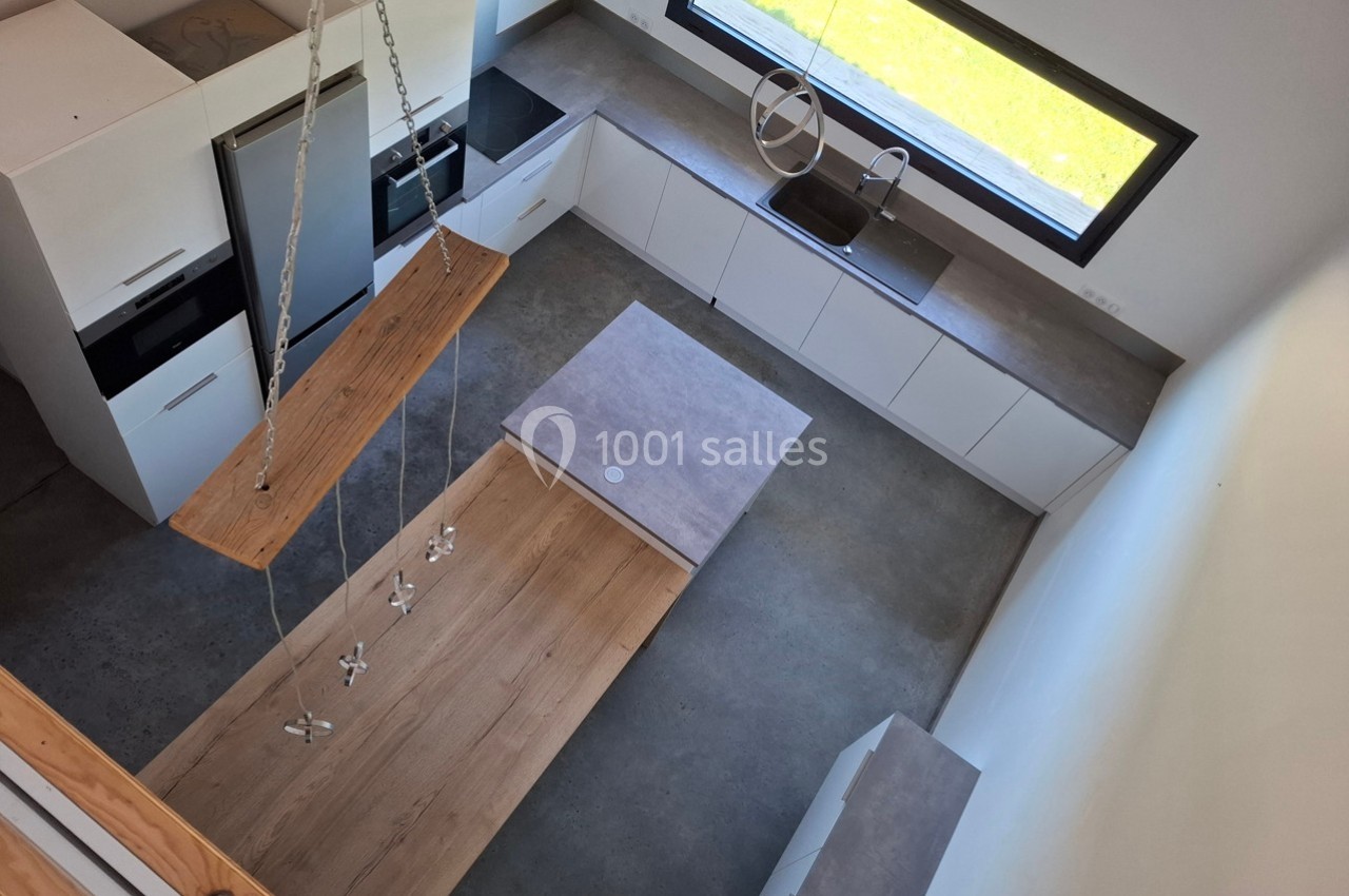 Vue d'une cuisine moderne avec îlot central, plan de travail en béton, meubles blancs et grande fenêtre.