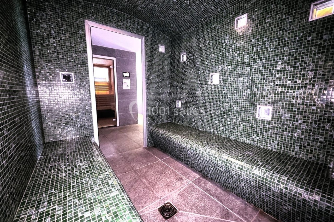 Hammam aux murs et bancs recouverts de mosaïques vertes, avec une porte ouverte menant à une autre pièce.
