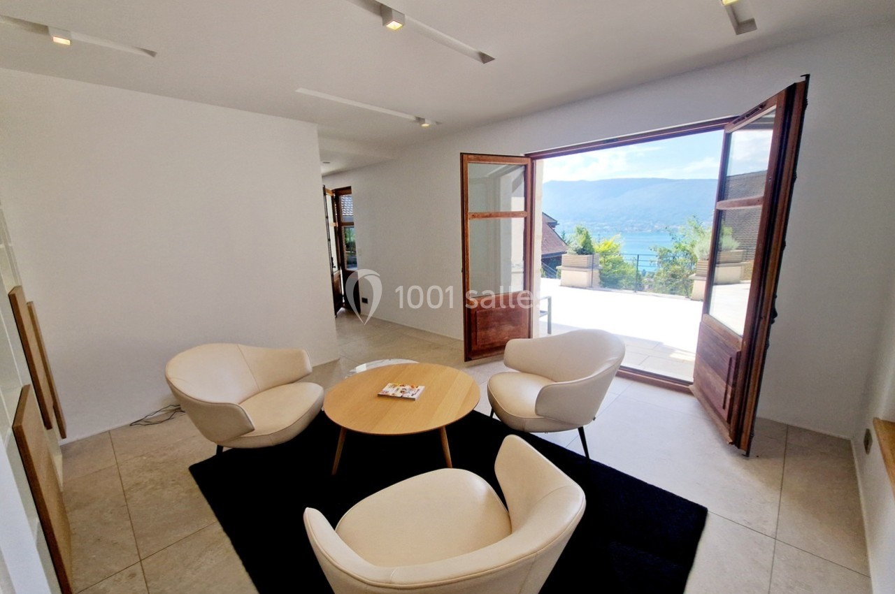 Salon lumineux avec quatre fauteuils autour d'une table basse, ouvert sur une terrasse avec vue sur un lac.