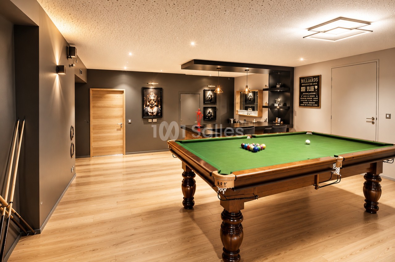 Salle de jeux avec billard au tapis vert, bar noir avec rangements éclairés, murs gris et beige, sol en bois clair.