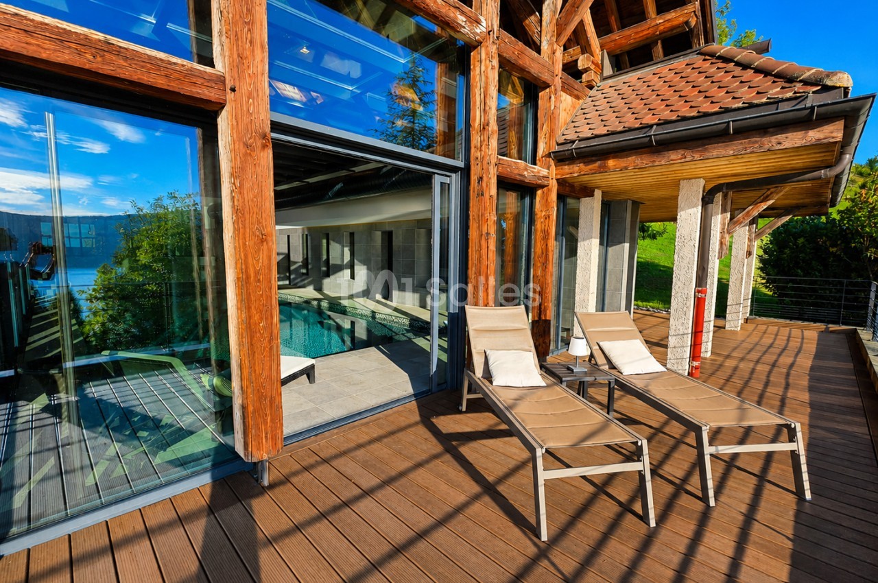 Terrasse en bois avec chaises longues face à baies vitrées donnant sur piscine et lac.