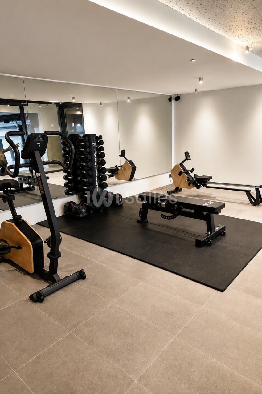 Salle de fitness équipée de vélos, rameurs et haltères, sol gris, miroir mural et éclairage indirect.