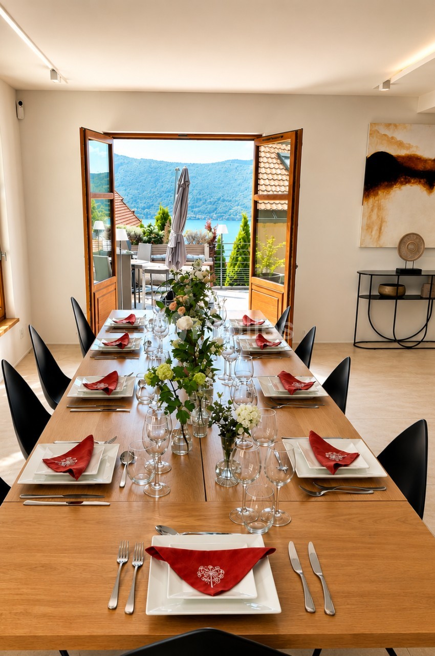 Salle à manger dressée avec table en bois, chaises noires, serviettes rouges et vue sur lac de montagne par portes ouvertes.
