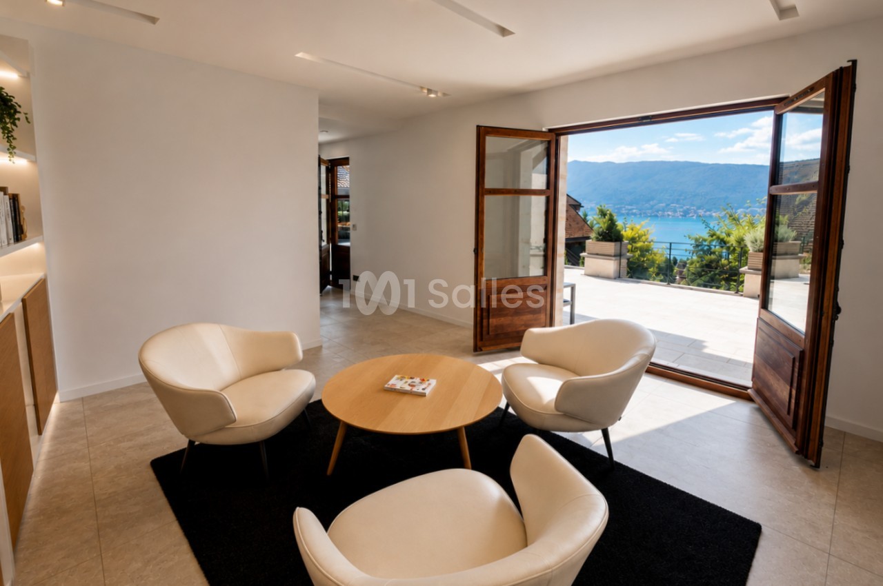 Salle intérieure moderne avec fauteuils beige, table bois rond et portes ouvertes sur terrasse avec vue lac.