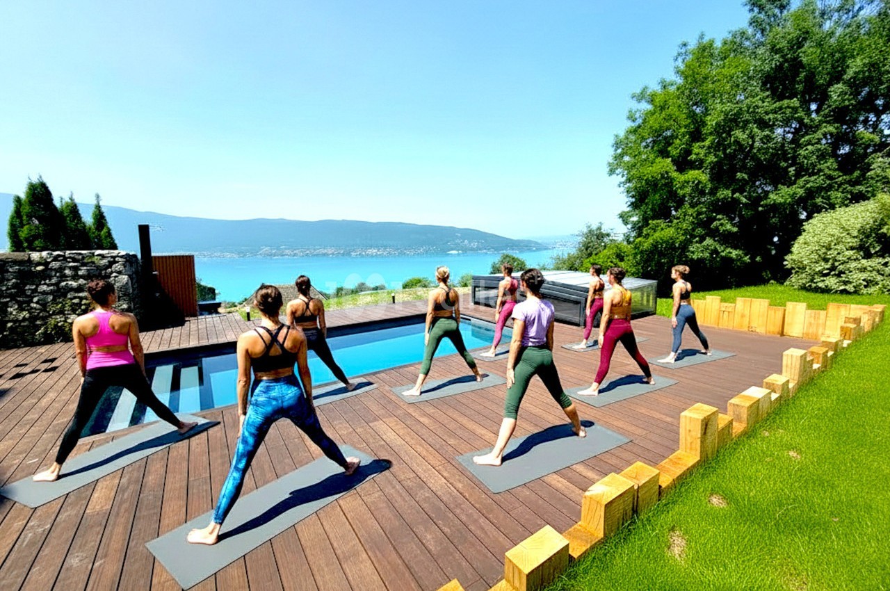 Un groupe de personnes pratique le yoga en plein air sur une terrasse en bois près d'une piscine avec vue sur un lac.