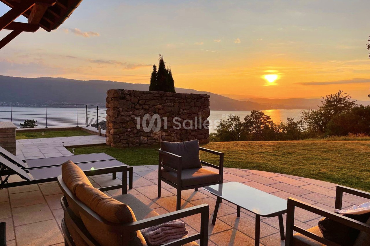 Terrasse avec mobilier de jardin, vue sur un lac au coucher du soleil, entourée de verdure et d'un mur en pierre.