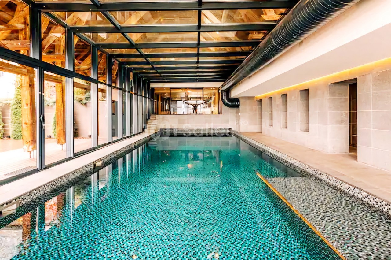 Piscine intérieure avec carrelage mosaïque, entourée de murs en pierre et d'une structure en verre et bois.