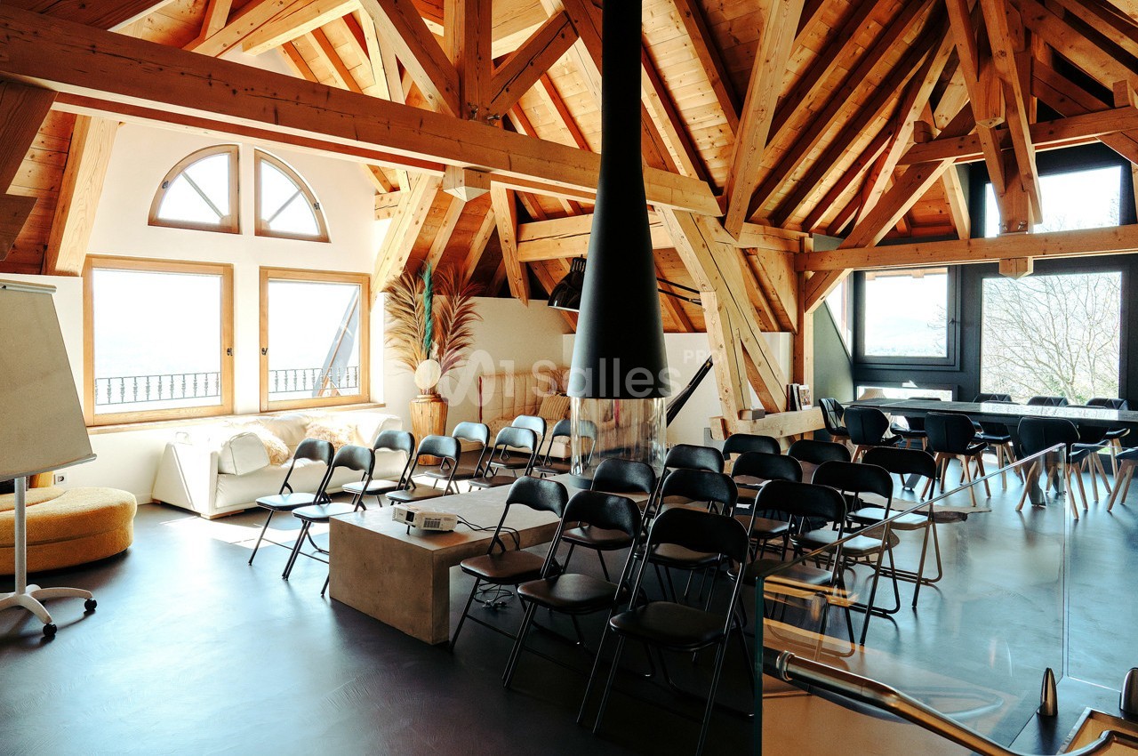 Salle lumineuse avec charpente en bois apparente, aménagée avec des chaises noires disposées face à un tableau blanc.