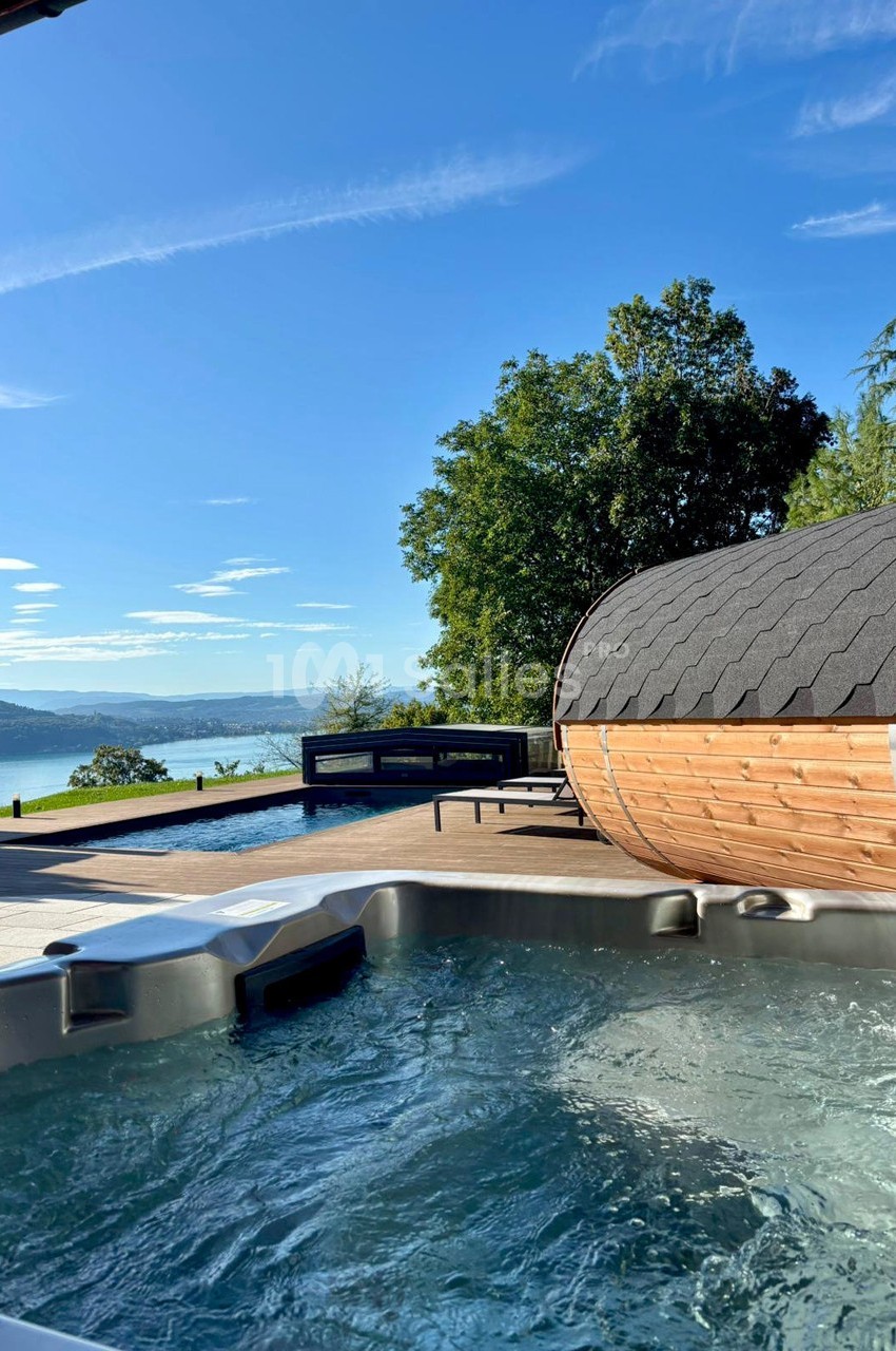 Vue sur un jacuzzi, une piscine, un sauna en bois et un paysage de lac entouré de collines sous un ciel bleu.