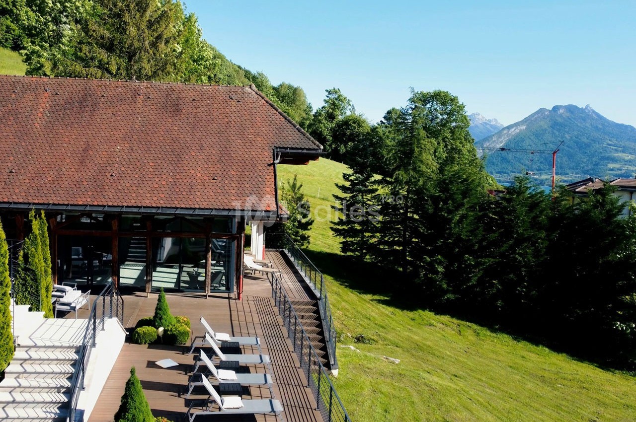 Terrasse en bois avec chaises longues, bordée d'arbres, donnant sur des collines verdoyantes et des montagnes au loin.