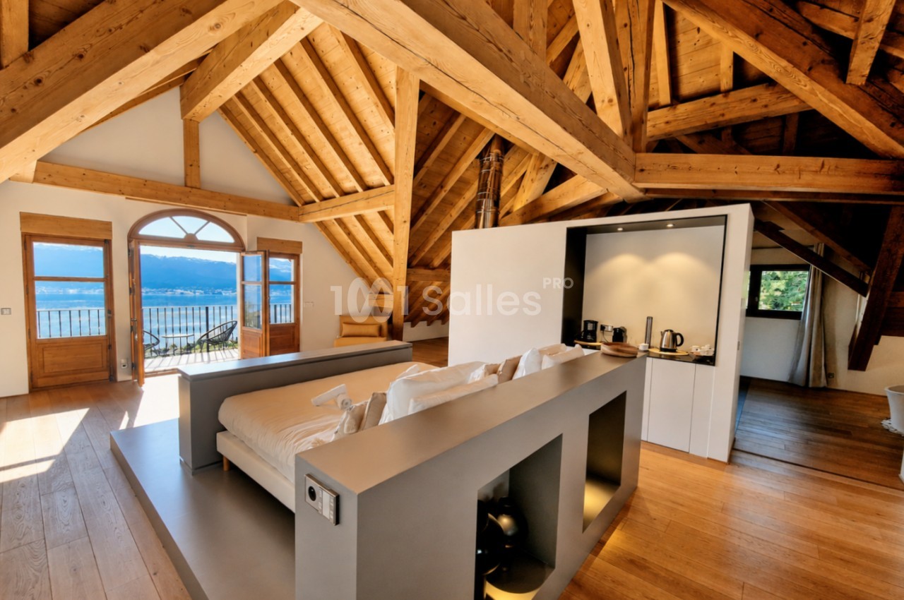 Loft mansardé avec poutres apparentes, canapé central, vue lac par baies vitrées et balcon.