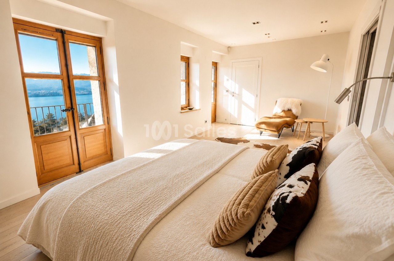 Chambre spacieuse avec lit double, portes-fenêtres en bois ouvrant sur balcon avec vue lac, mobilier épuré.