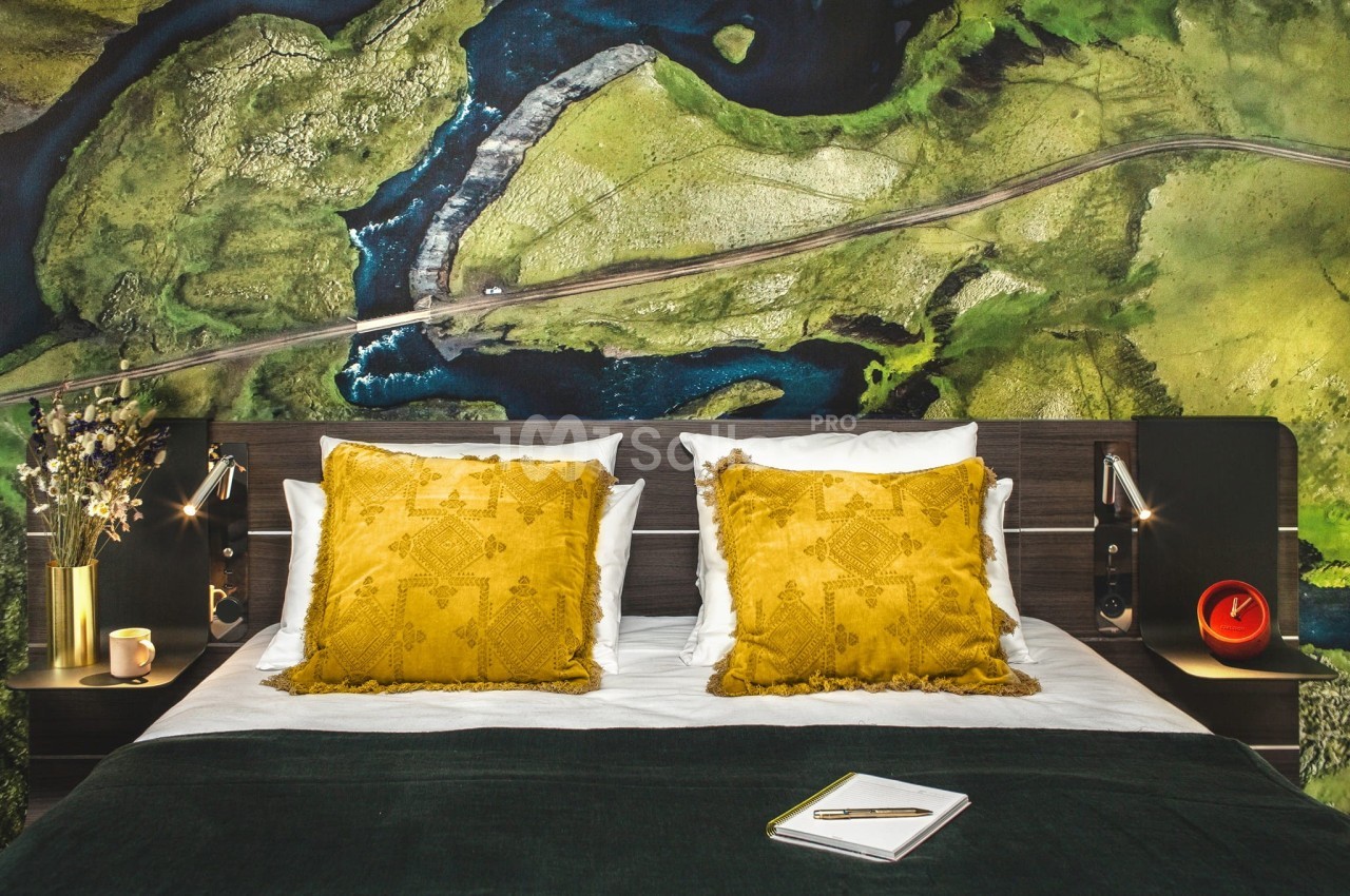 Lit double avec coussins jaunes, couverture verte et décoration murale représentant une vue aérienne de paysage naturel.