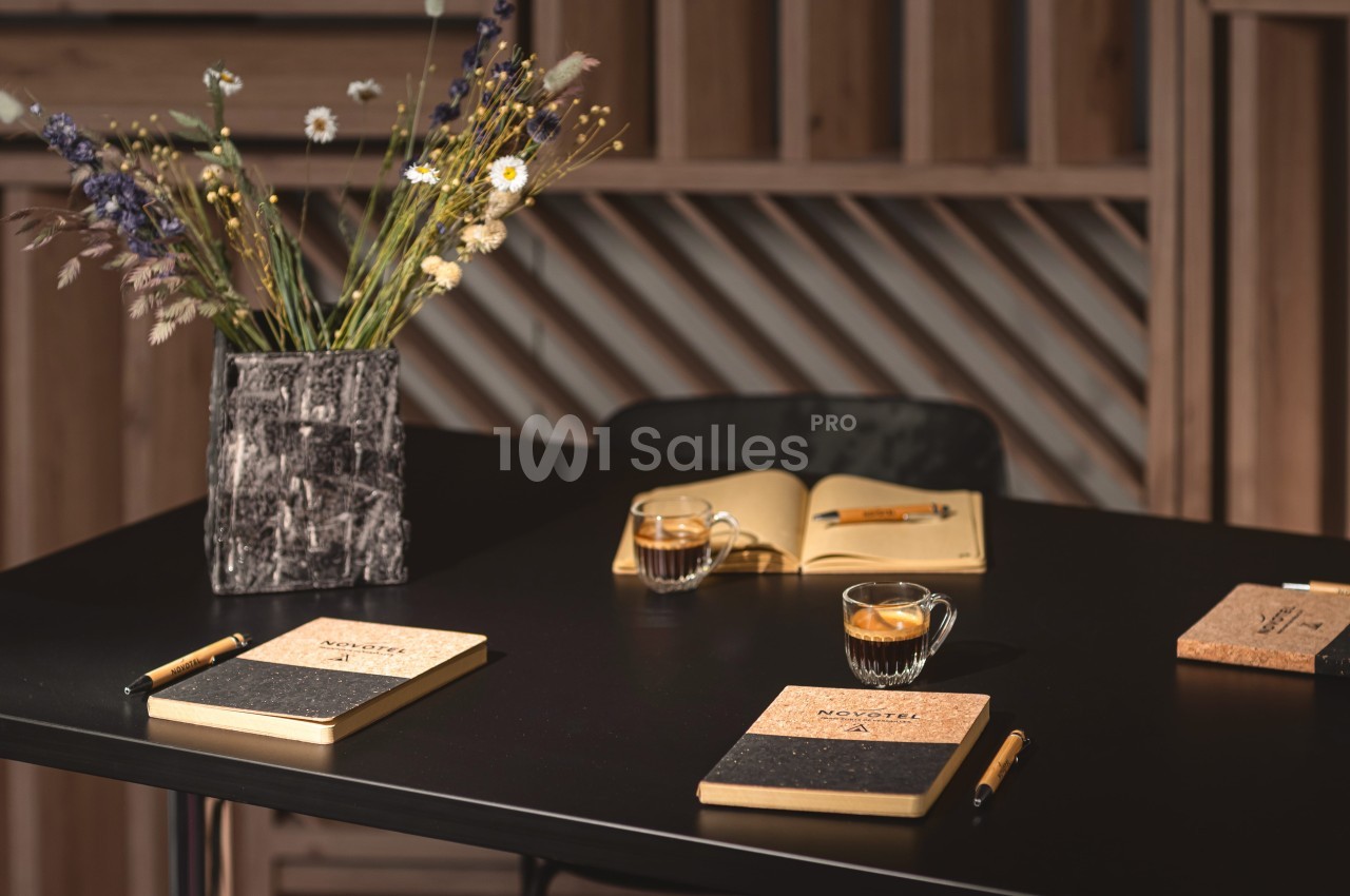 Carnet et stylos sur une table noire avec des tasses de café, un vase de fleurs séchées et un carnet ouvert en arrière-plan.