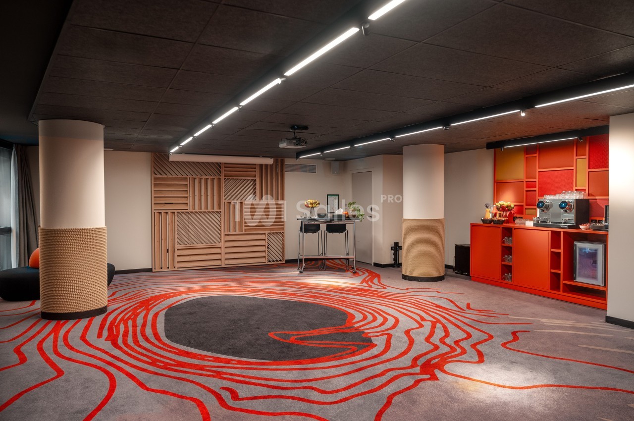 Salle moderne avec tapis aux motifs rouges, colonnes blanches, éclairage au plafond et coin café équipé à droite.