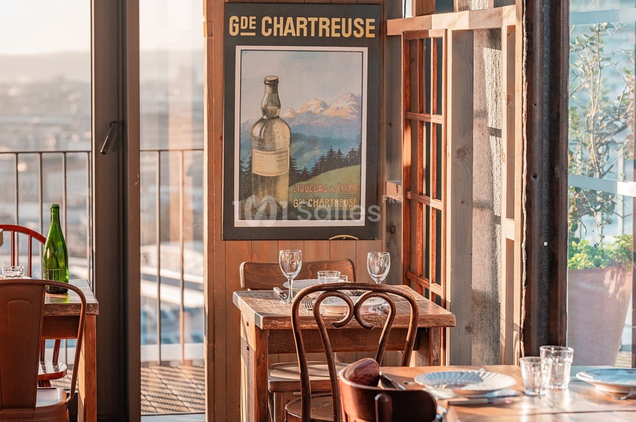 Salle de restaurant lumineuse avec des tables en bois, une affiche de la Chartreuse et une vue sur l'extérieur.