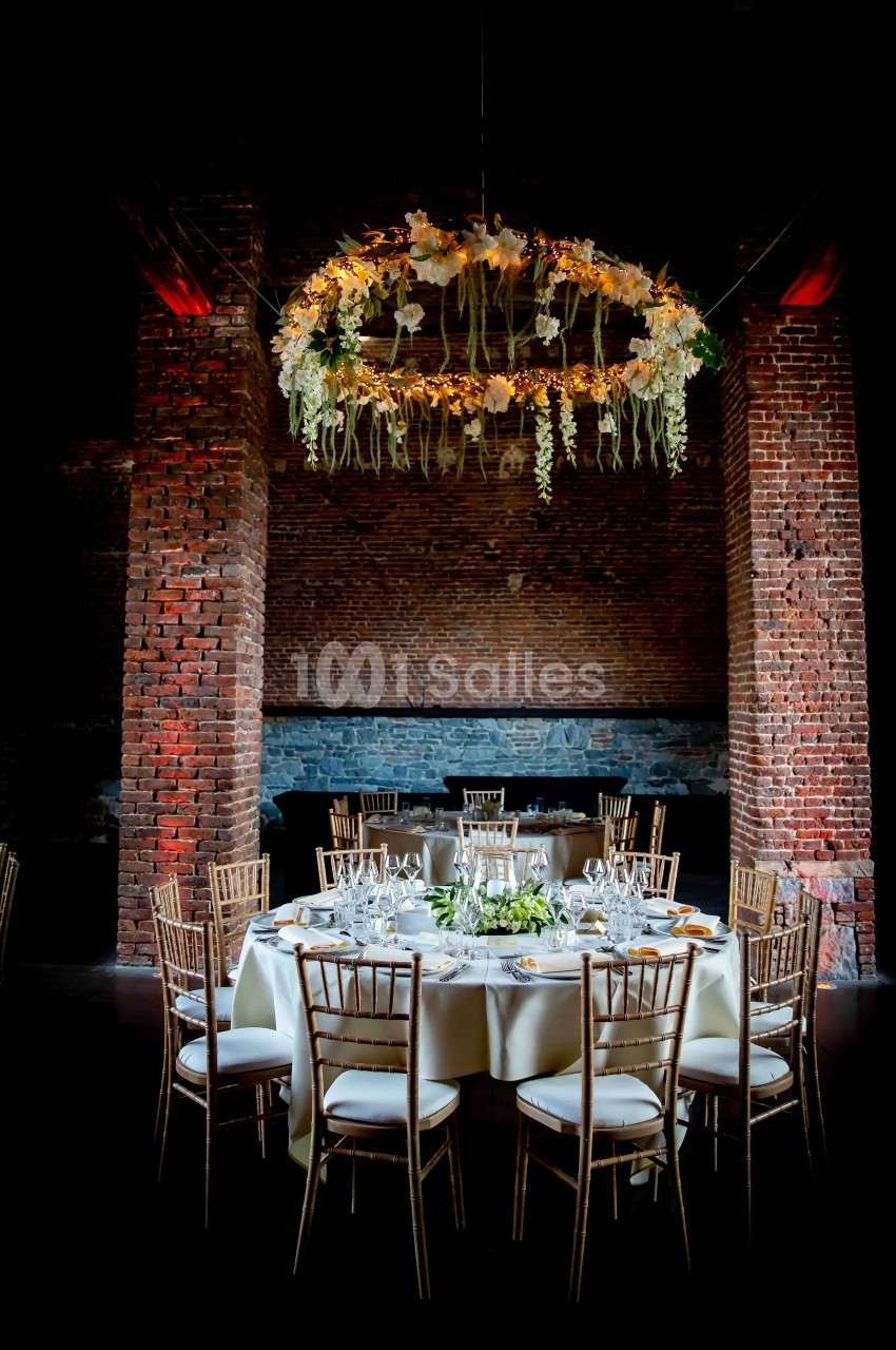 Table ronde élégamment dressée avec des chaises en bois, sous un lustre floral suspendu dans une salle en briques.