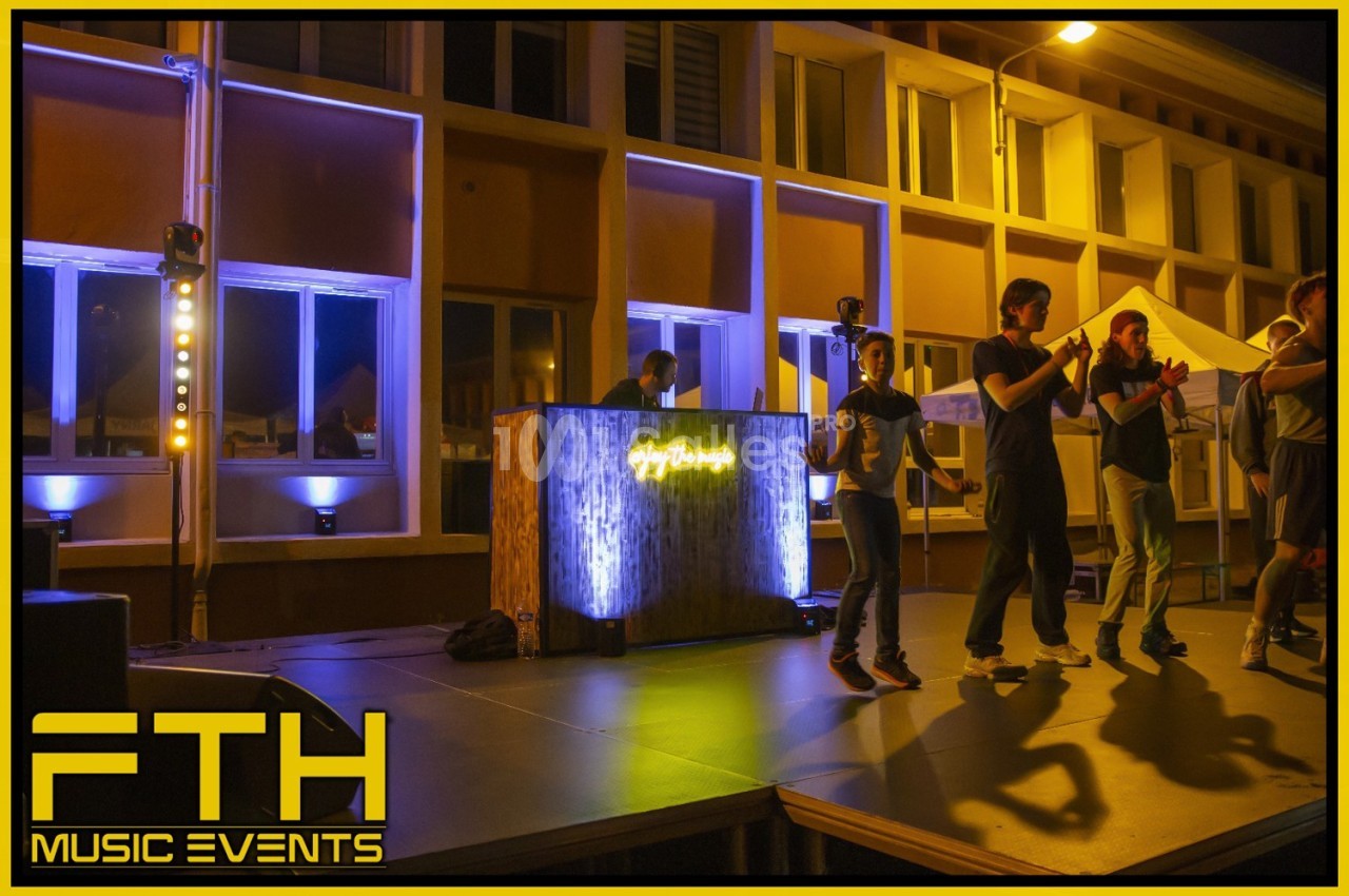 Des personnes sur une scène éclairée applaudissent près d'une table de DJ lumineuse, en extérieur de nuit.