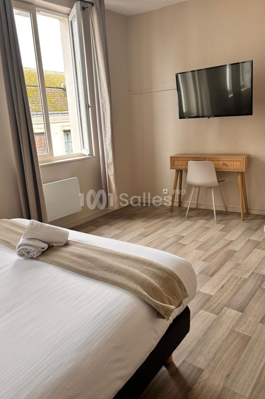 Chambre lumineuse avec lit double, bureau en bois, chaise blanche, télévision murale et fenêtre donnant sur des toits.